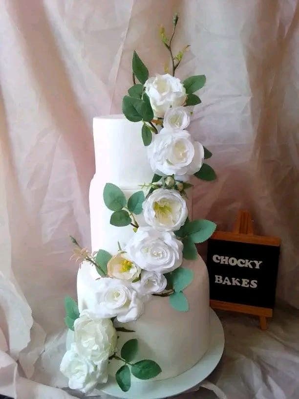 Three-tier white cake with a cascade of white roses, beside a small Chockybakes sign.