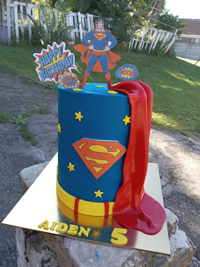 Blue themed children's cake with a red cape, yellow stars and a superhero topper.